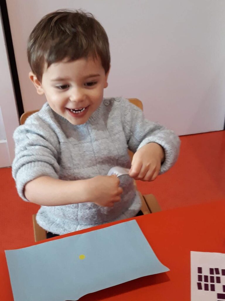 PHOTO D'UN PETIT GARÇON ENTRAIN DE SOURIRE ET DE COLLER DES PASTILLES SUR UNE FEUILLE